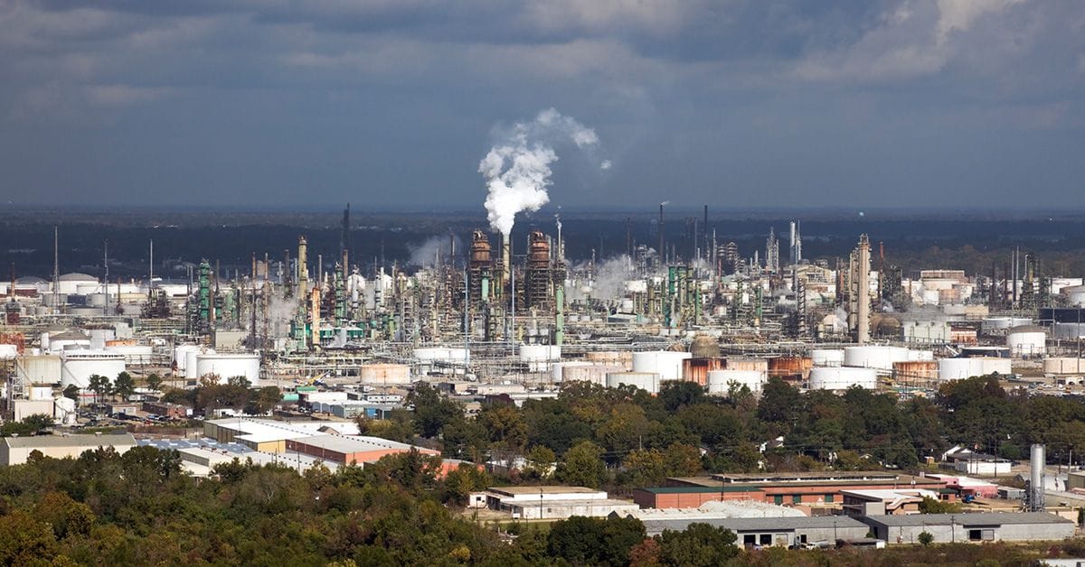 A large industrial refinery complex with multiple smokestacks emitting white smoke, surrounded by various buildings and green areas, under a partly cloudy sky.