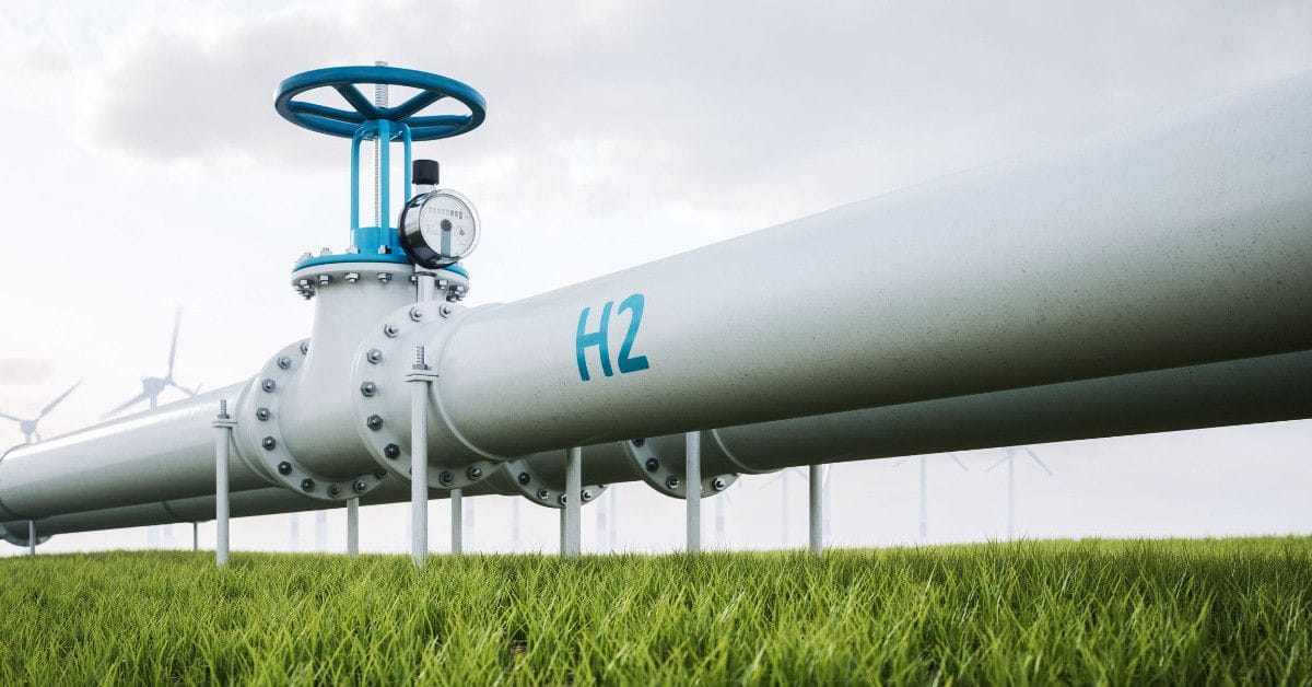 Industrial hydrogen pipeline labeled “H2” with a large valve and gauge, extending across a grassy field with multiple wind turbines under a cloudy sky.