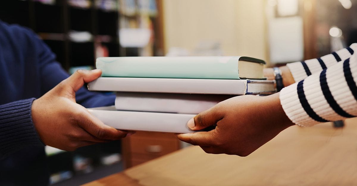 A Close‑up image showing a stack of books being passed between two people across a desk in an indoor setting.