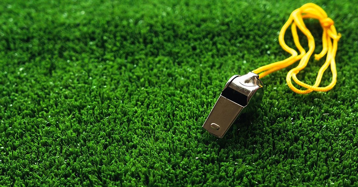 A metal sports whistle with a yellow string resting on a green, grassy soccer field.