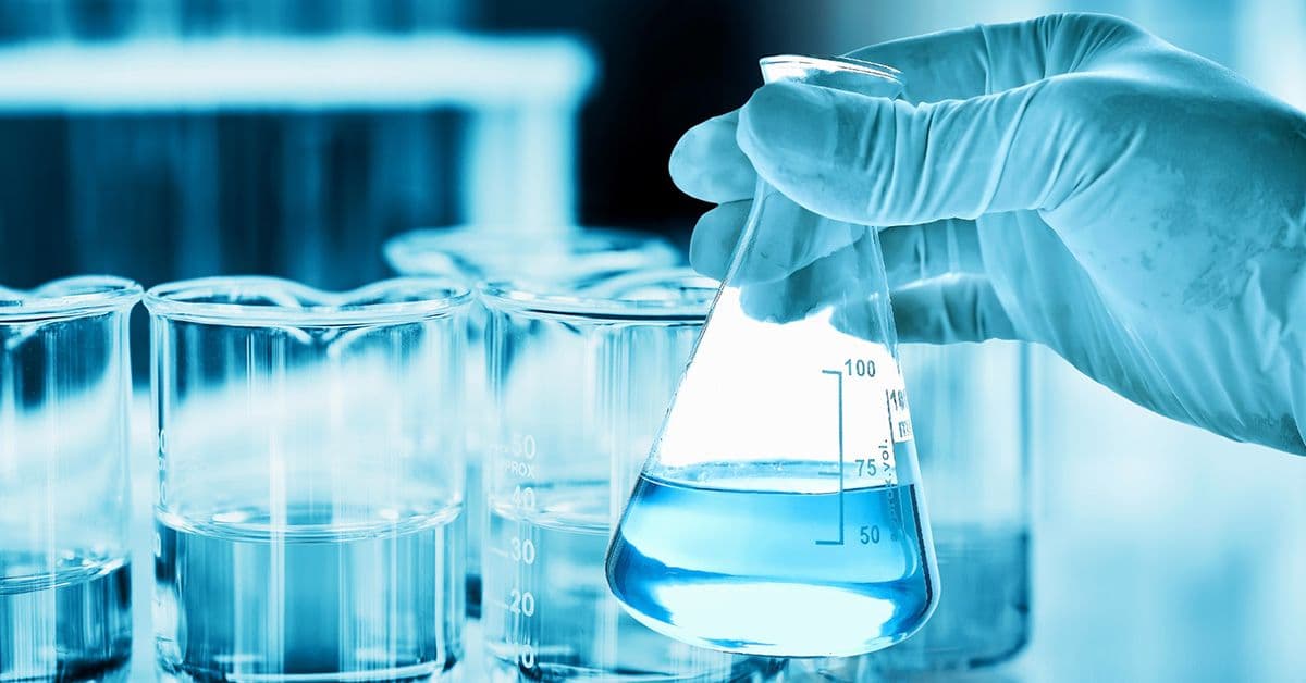 A scientist's gloved hand holding a beaker of blue liquid in a chemistry lab, with other beakers and vials in the background.