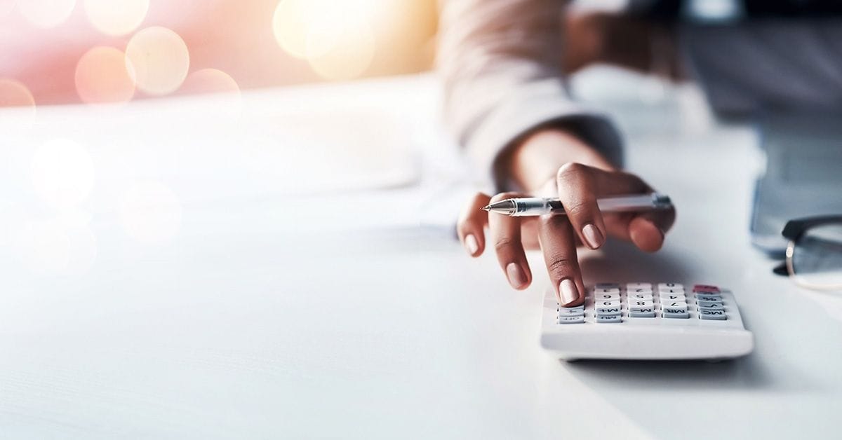 A person uses a calculator while holding a pen, with a blurred background featuring circular light flares.