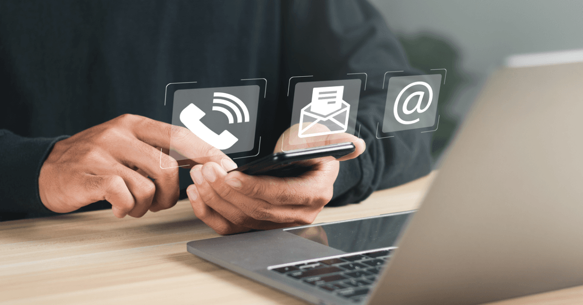 A person holding a smartphone, interacting with icons for phone, email, and messaging, beside a laptop on a wooden desk.