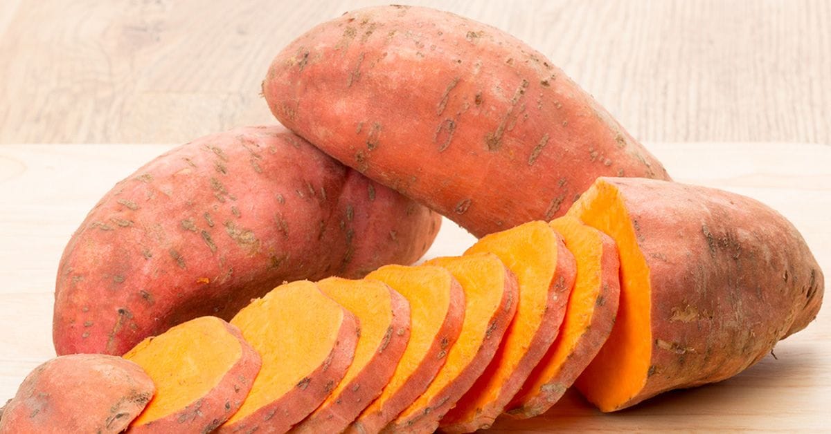 Two whole sweet potatoes and one partially sliced sweet potato with bright orange flesh arranged on a light wooden surface.