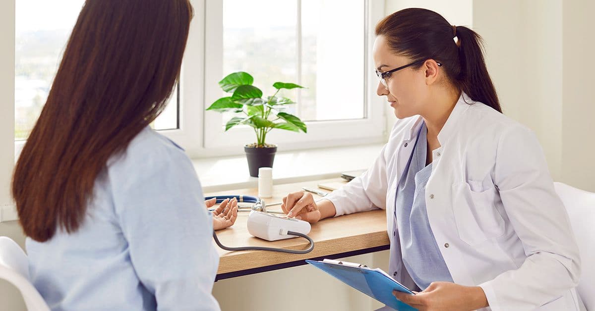 A healthcare professional in a white coat measures a patient's blood pressure in a bright office, surrounded by plants and medical equipment.