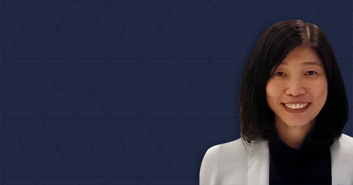 A woman with shoulder-length black hair smiles at the camera, standing against a dark blue geometric background. She is wearing a light-colored blazer over a dark top.