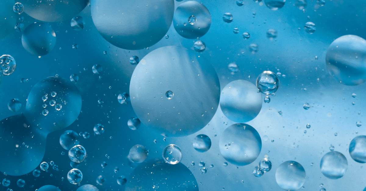 Macro photograph of round oil droplets in shades of blue, ranging from large spheres to tiny bubbles, suspended in water.