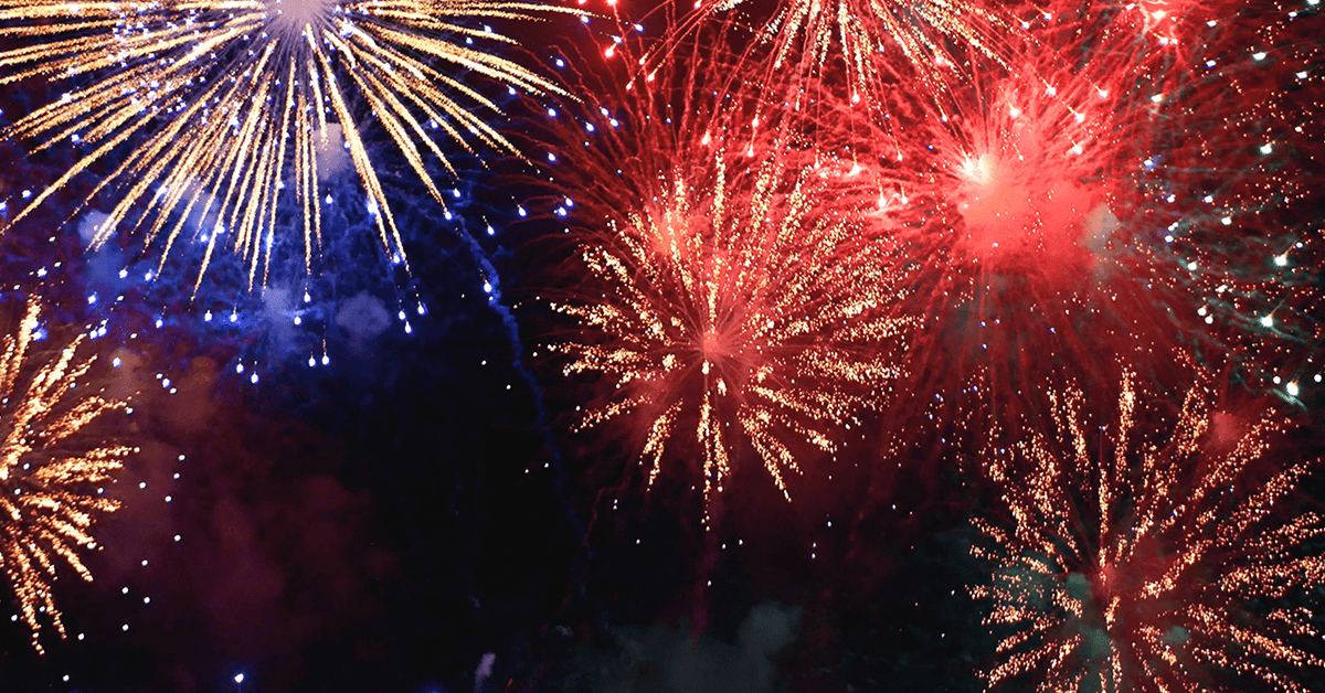 A vivid display of multiple fireworks in red, gold, and blue colors bursting across the night sky, with bright streaks and lingering smoke clouds.