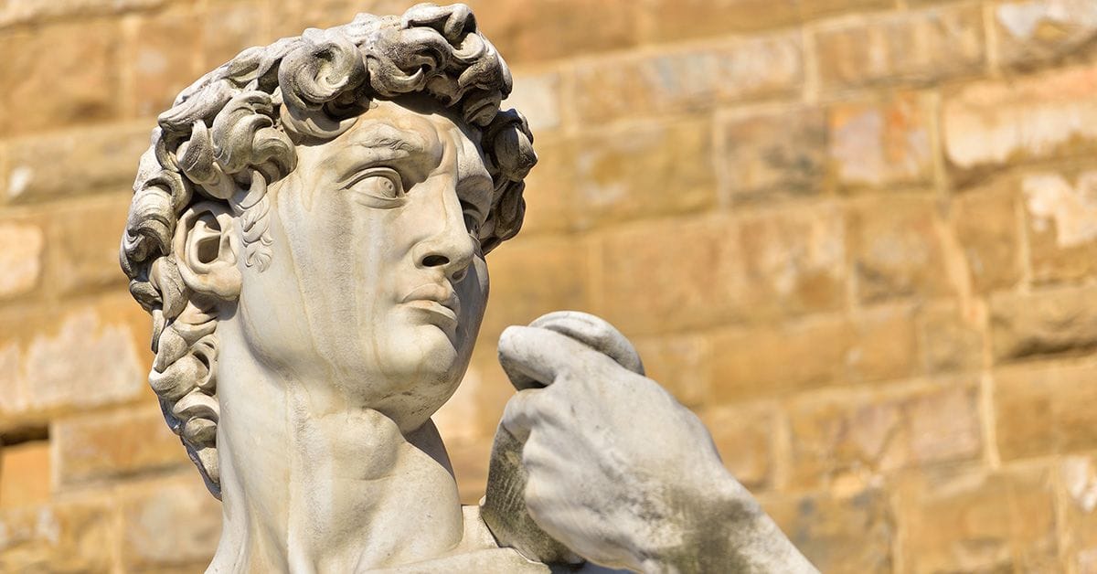 Close-up of a marble statue depicting a young man with curly hair, focusing on his detailed face and hand against a blurred stone wall background.