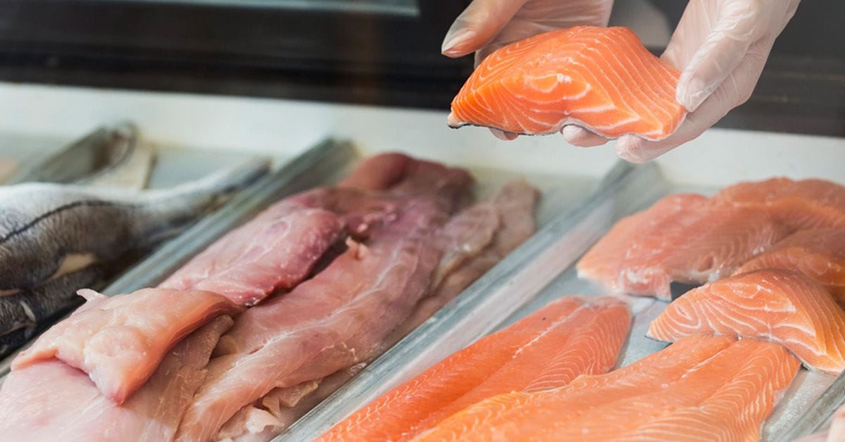 A person wearing disposable gloves is holding a fresh salmon fillet over a display case filled with various cuts of raw fish, including salmon and white fish fillets.