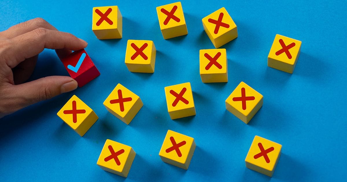 A hand is picking up a single red cube with a blue checkmark from a scattered group of yellow cubes marked with red Xs on a bright blue background.