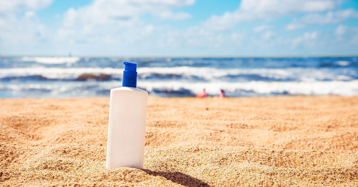 a generic, blank white bottle of sunscreen sitting on a sunny, sandy beach with ocean waves in the background
