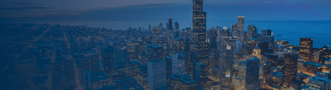 Aerial view of a city skyline with tall skyscrapers, illuminated buildings, and a body of water at dusk.