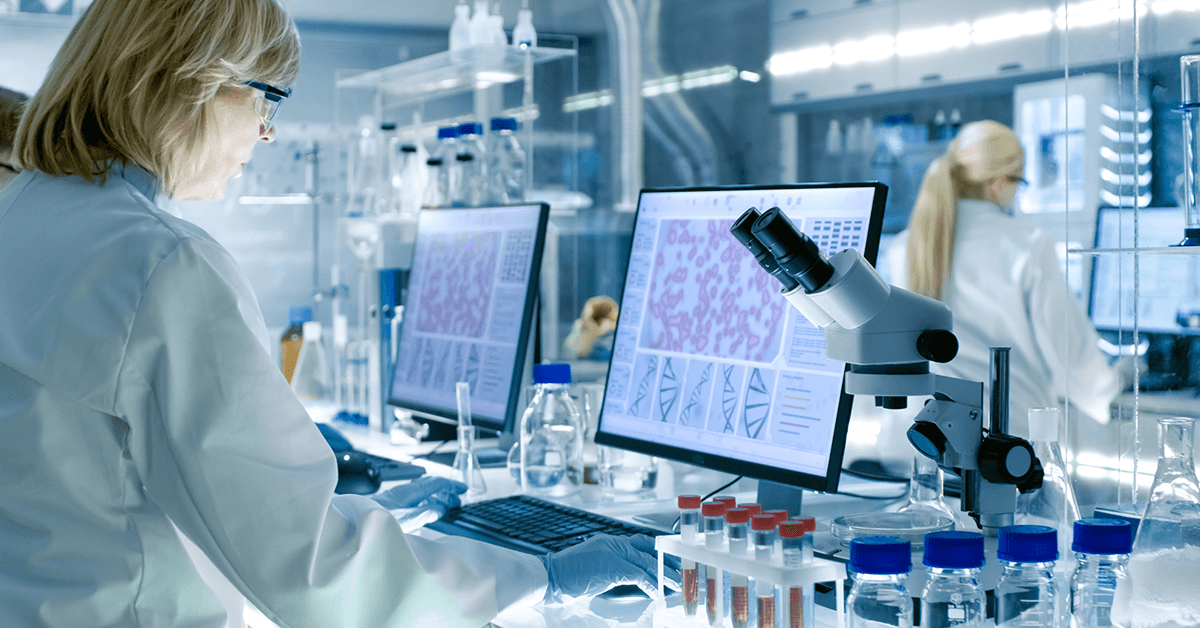 Laboratory scene showing a researcher wearing protective gloves and goggles, analyzing microscope images on computer screens with vials, flasks, and a microscope on the bench