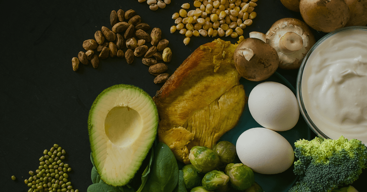 A flat lay of nutritious foods including avocado, eggs, chicken, broccoli, spinach, mushrooms, nuts, and yogurt on a dark background.