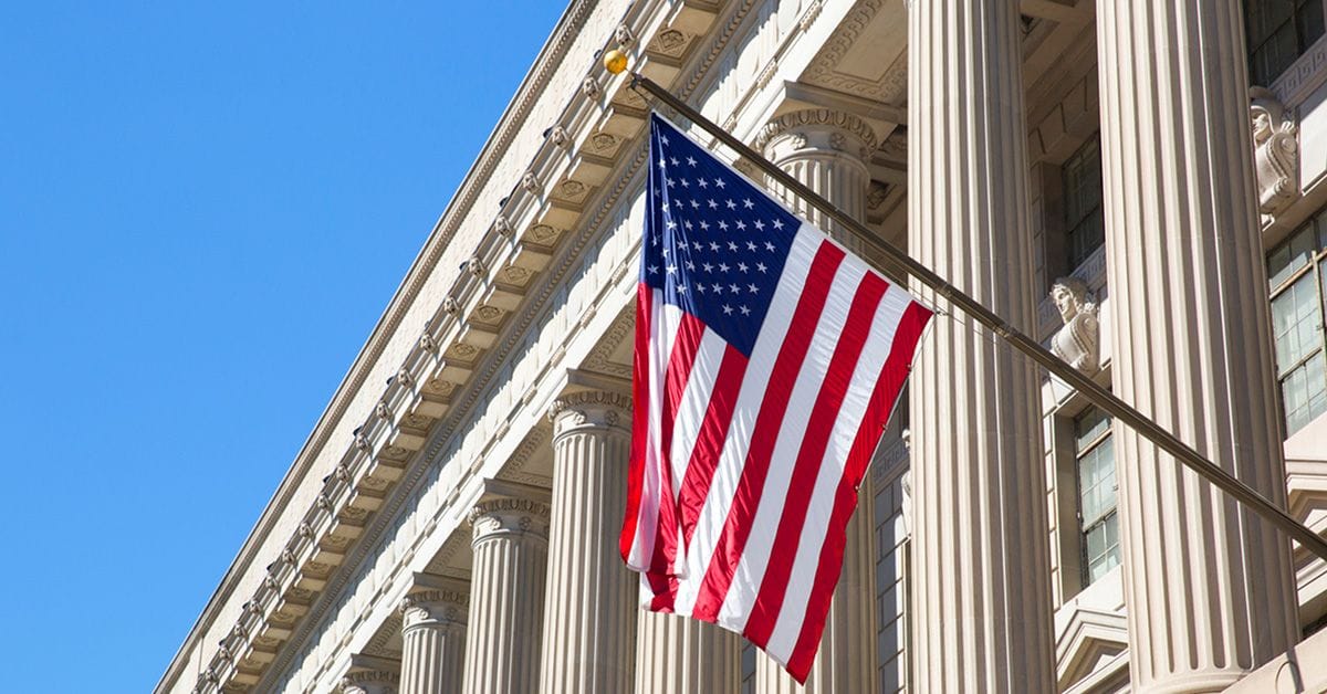 An American flag hangs from a pole attached to the façade of a large neoclassical building with tall columns, set against a clear blue sky.