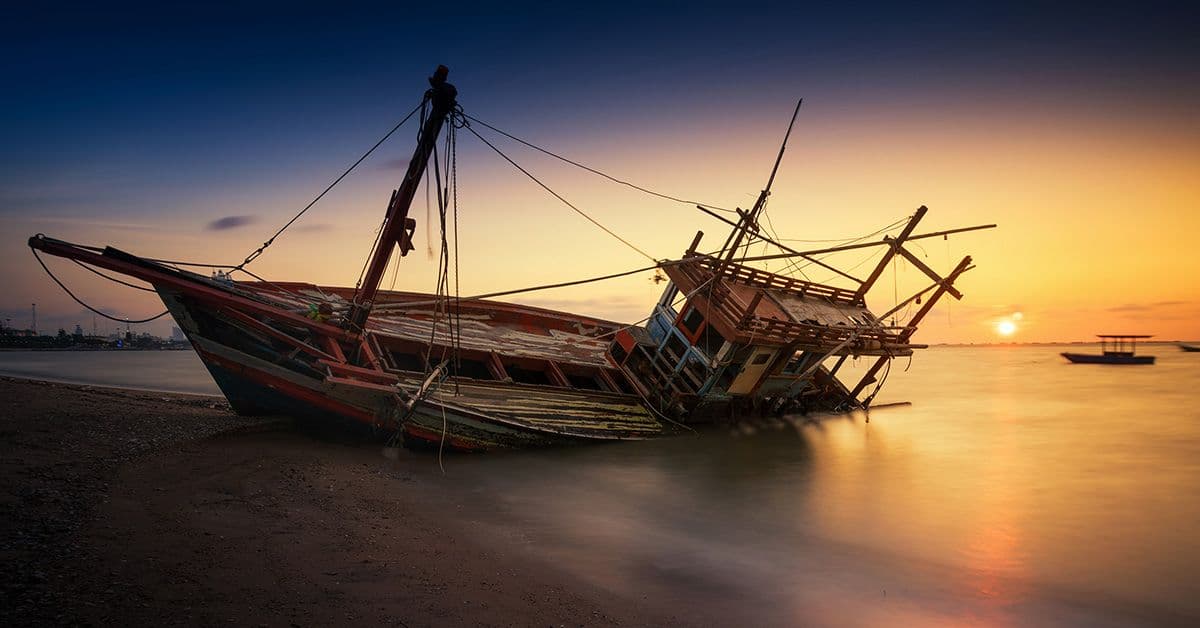 a small wooden shipwrecked boat on the shoreline with a setting sun on the horizon