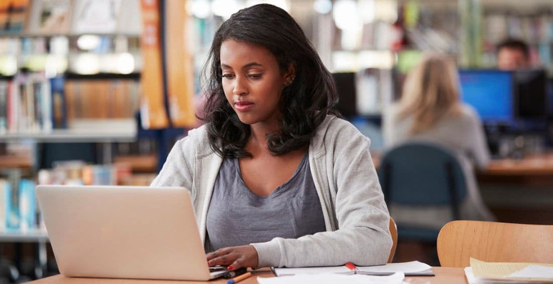 Woman working on a laptop in a library, surrounded by books and papers.