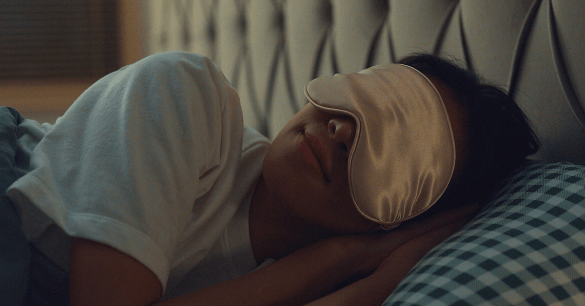 Close-up of a person lying on their side in bed with hands under their head, wearing a satin sleep mask and resting on a patterned pillow.