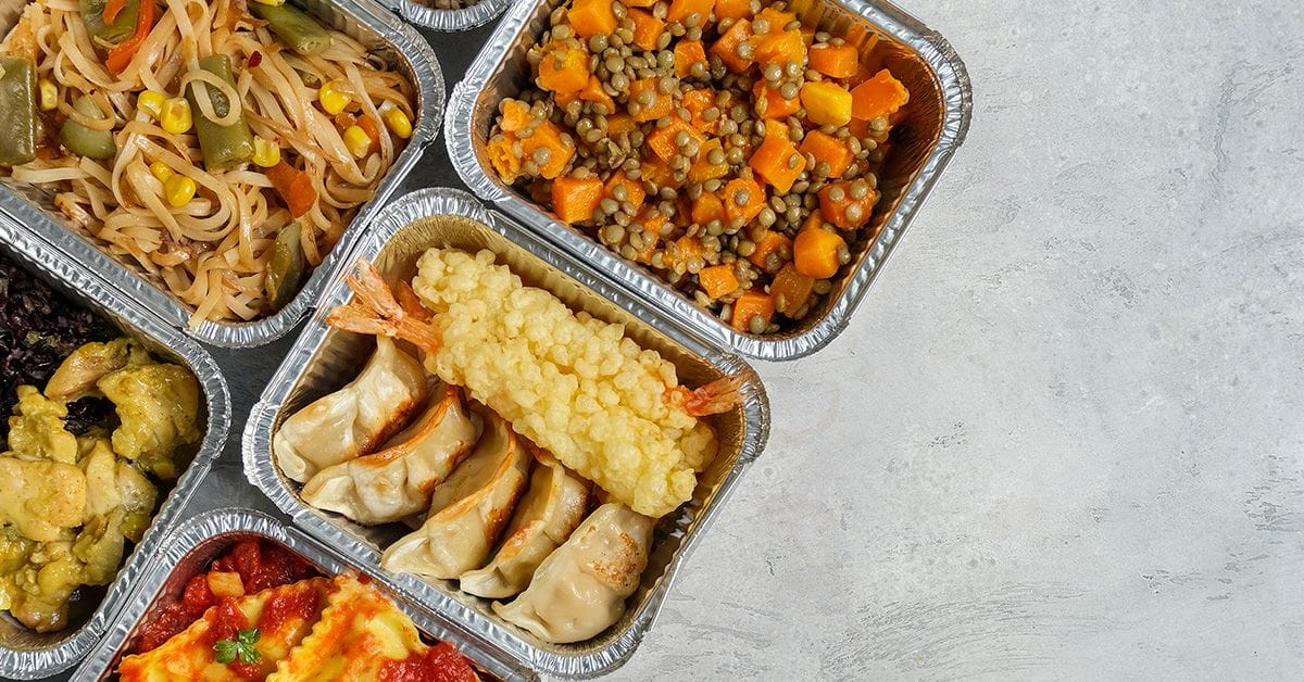 A variety of meals in foil containers, including pasta, vegetable medley, dumplings, tempura shrimp, and baked dishes, arranged on a light gray surface.