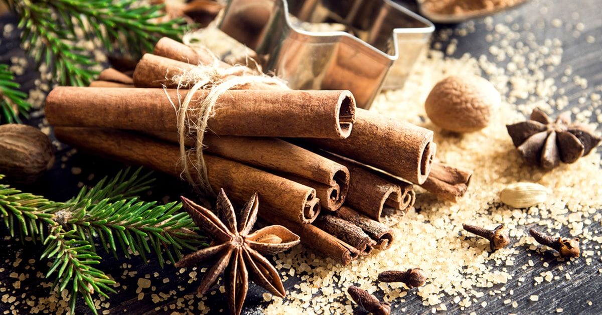 A bundle of cinnamon sticks tied with twine surrounded by star anise, nutmeg, cloves, and coarse brown sugar on a dark wooden surface with evergreen sprigs and a metal cookie cutter.