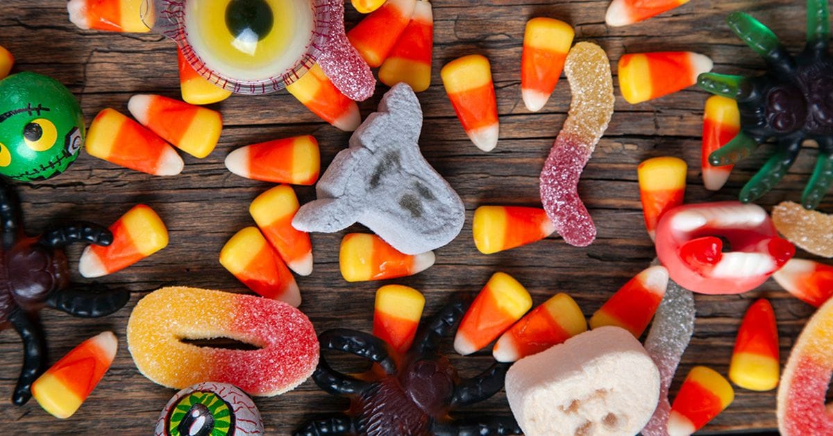 An assortment of halloween candies on a wooden table, including candy corn, gummy worms, gummy spiders, chocolate eyeballs, and marshmallow ghosts.