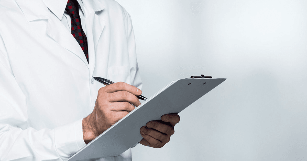 Close-up of a person wearing a lab coat and tie, holding a clipboard and writing with a pen against a plain background.