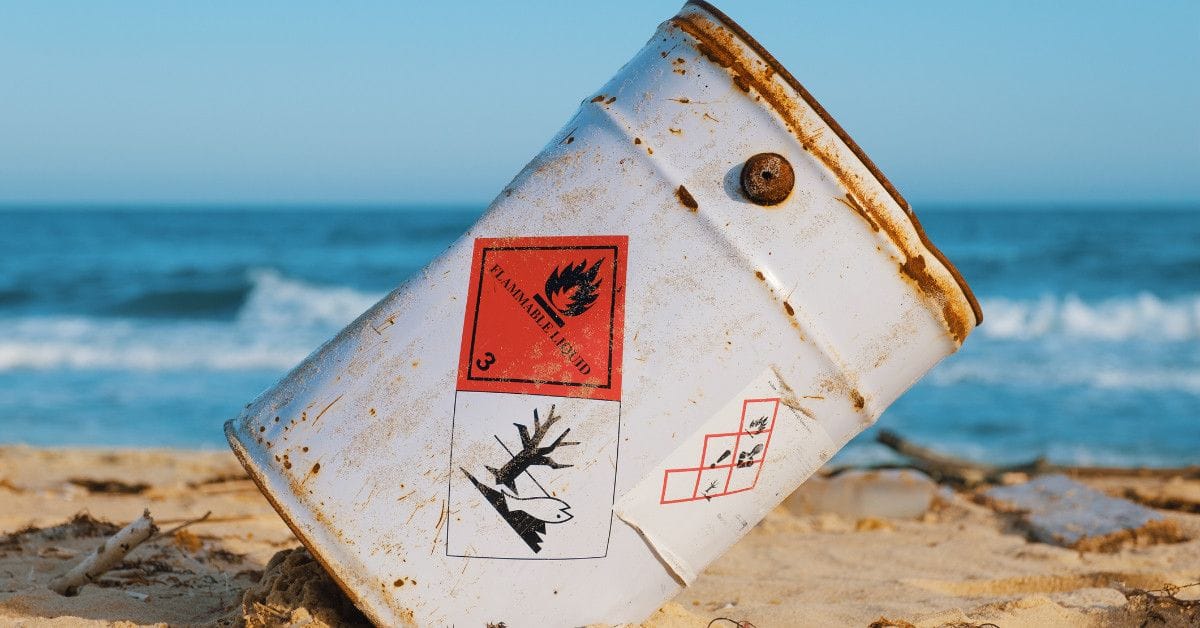 A Weathered metal drum displaying flammable liquid and environmental hazard symbols resting on a sandy beach near the ocean.