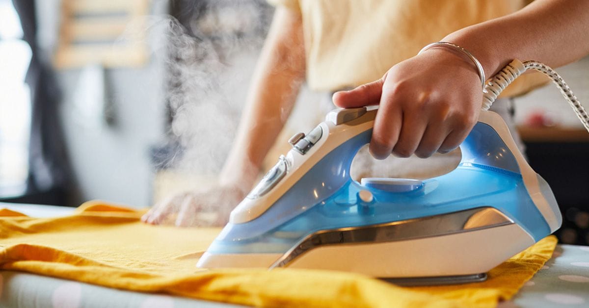 An individual is using a blue and white steam iron to press a yellow garment on an ironing board covered with a polka dot fabric, with visible steam rising from the iron in a bright indoor setting.