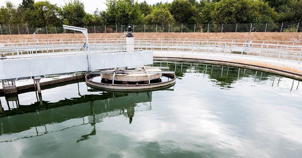 Wastewater treatment facility with a circular clarifier basin, surrounded by railings and trees in the background.