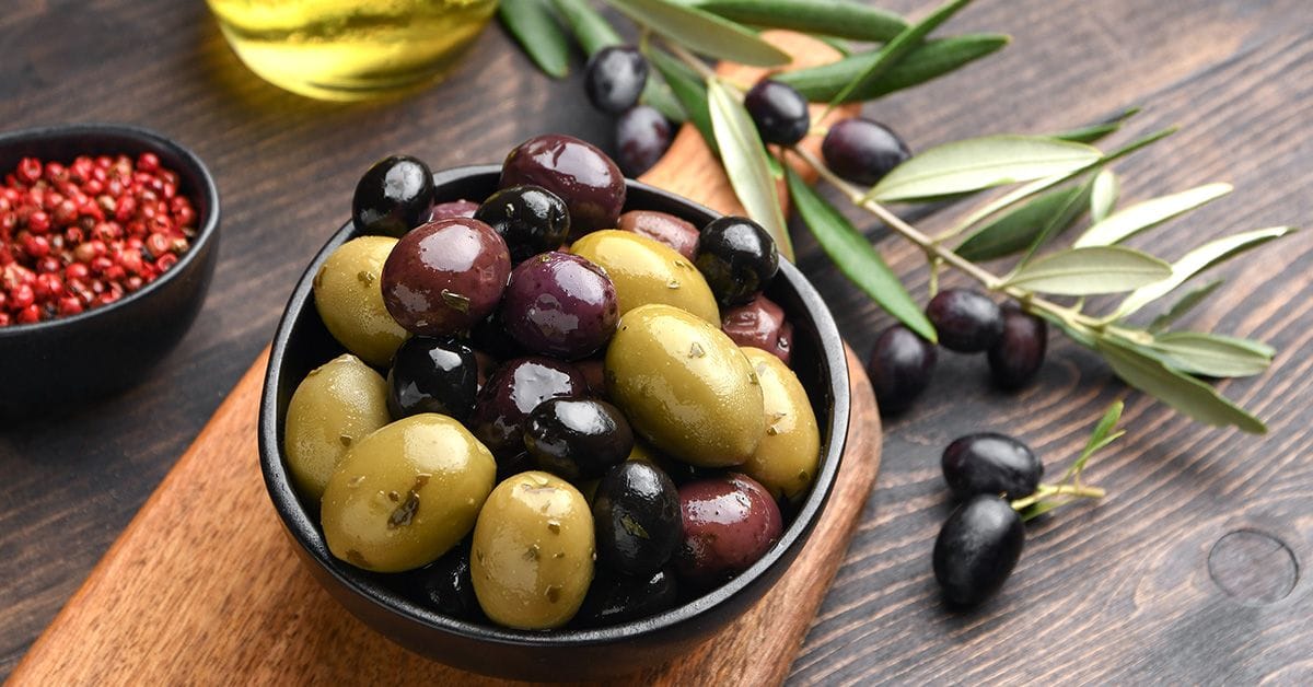 A bowl of mixed olives on a wooden board, with olive branches and a small bowl of red peppercorns in the background.