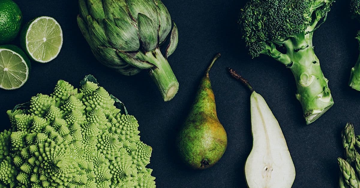 Various green vegetables and fruits, including artichoke, broccoli, romanesco, pear, and lime, are arranged on a dark background.