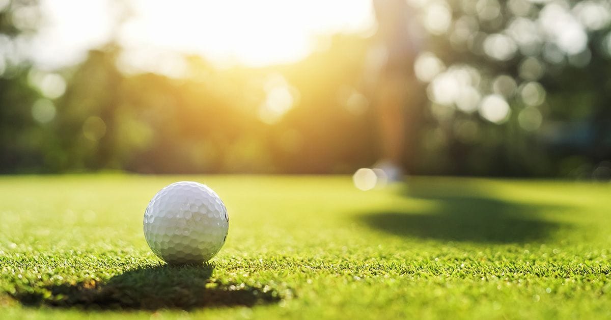 A close-up of a golf ball resting on green grass, with sunlight filtering through trees in the background, creating a warm atmosphere.