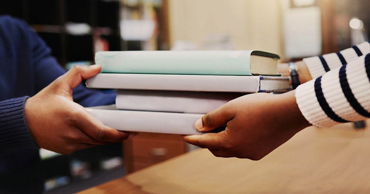 Two pairs of hands exchanging a stack of books, one wearing a striped sweater, the other showing a partial blue sleeve.