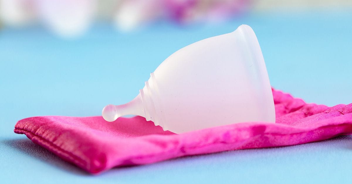 A close-up image of a translucent silicone menstrual cup resting on a bright pink cloth pouch against a light blue background.