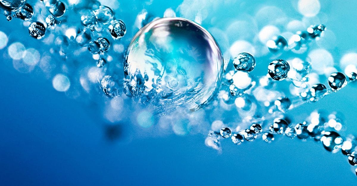 Close-up macro photograph of a translucent water droplet catching colorful light reflections, surrounded by streams of smaller water bubbles arranged diagonally across a gradient blue background.