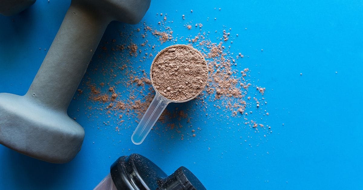 A dumbbell, a scoop of protein powder, and a shaker bottle on a blue surface.