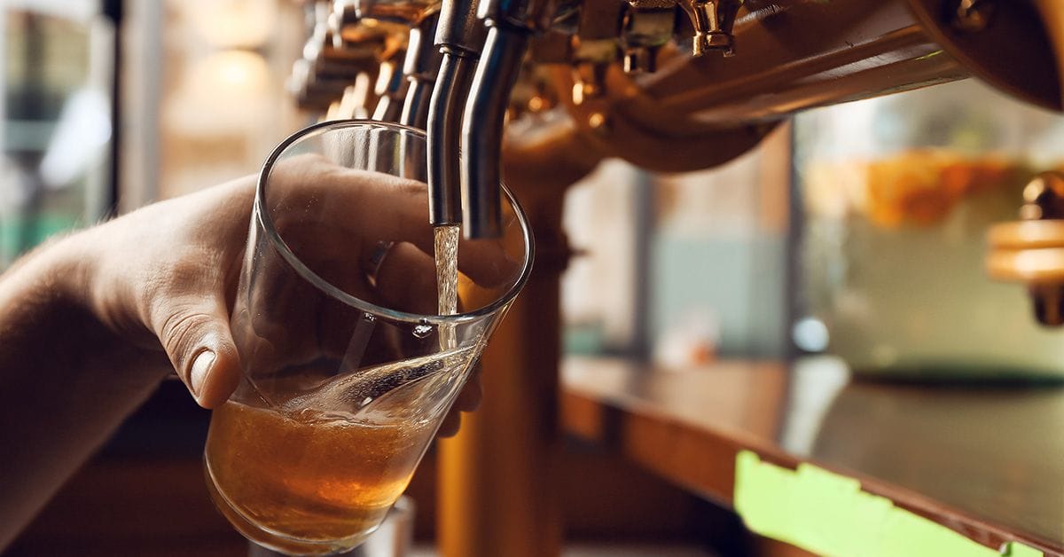A hand pours a golden beer into a glass from a tap, with blurred brewery equipment in the background.